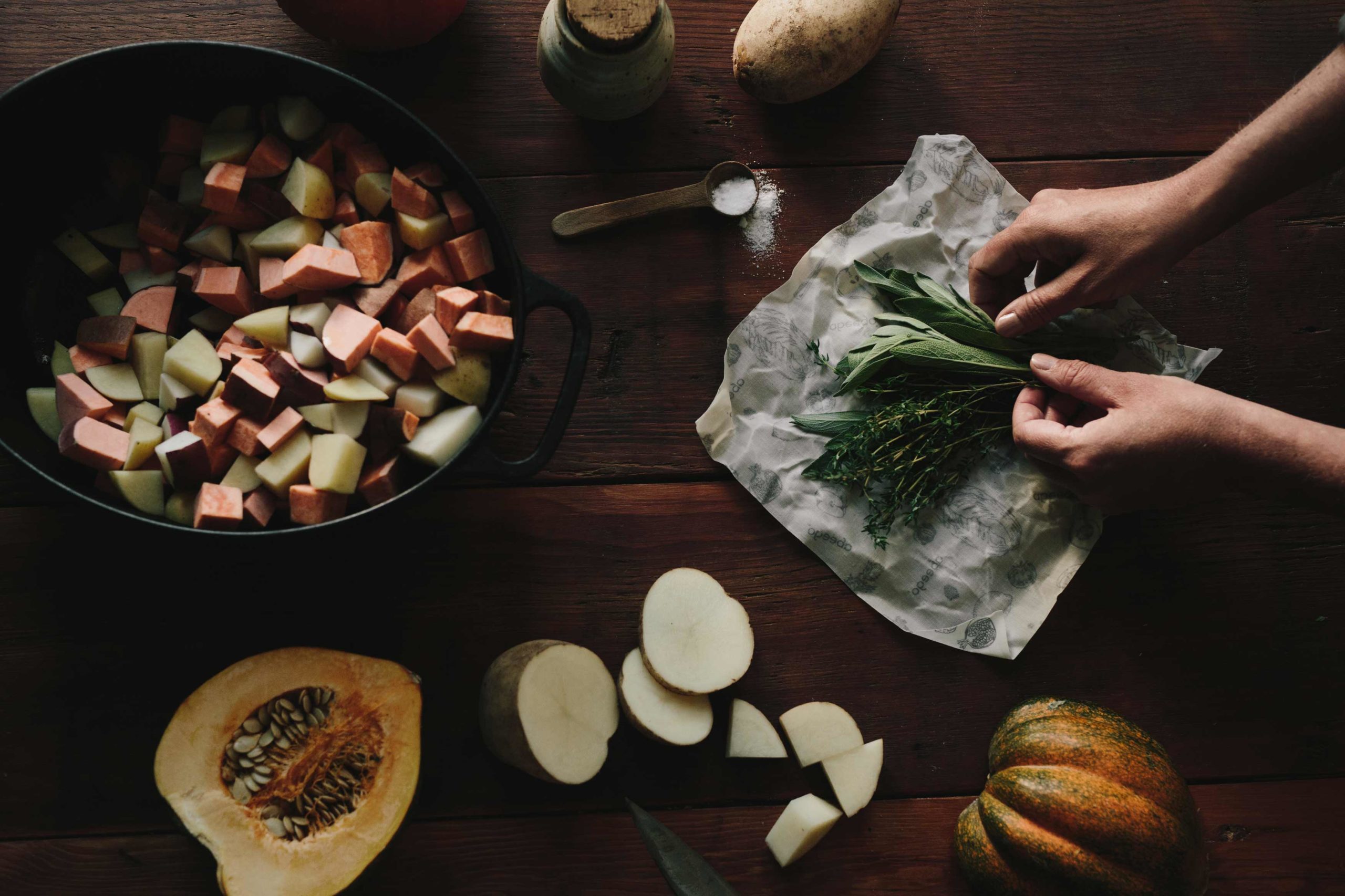 Coup de frais généraux d'une table en bois avec un grand pot en fonte avec des légumes-racines coupés, une demi-courge et des pommes de terre en tranches. Il y a un emballage ouvert d'emballage alimentaire en cire d'Abeego contenant des herbes