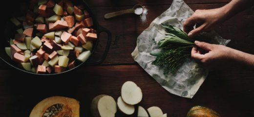 Overhead shot of a wooden table with a large cast iron pot with cut up root vegetables, half a squash, and sliced potatoes. There is an open package of Abeego wax food wrap containing herbs